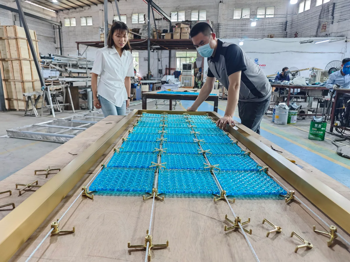 Two people beside a workbench holding blue textured crystal panels inside a gold-colored metal frame in a workshop.